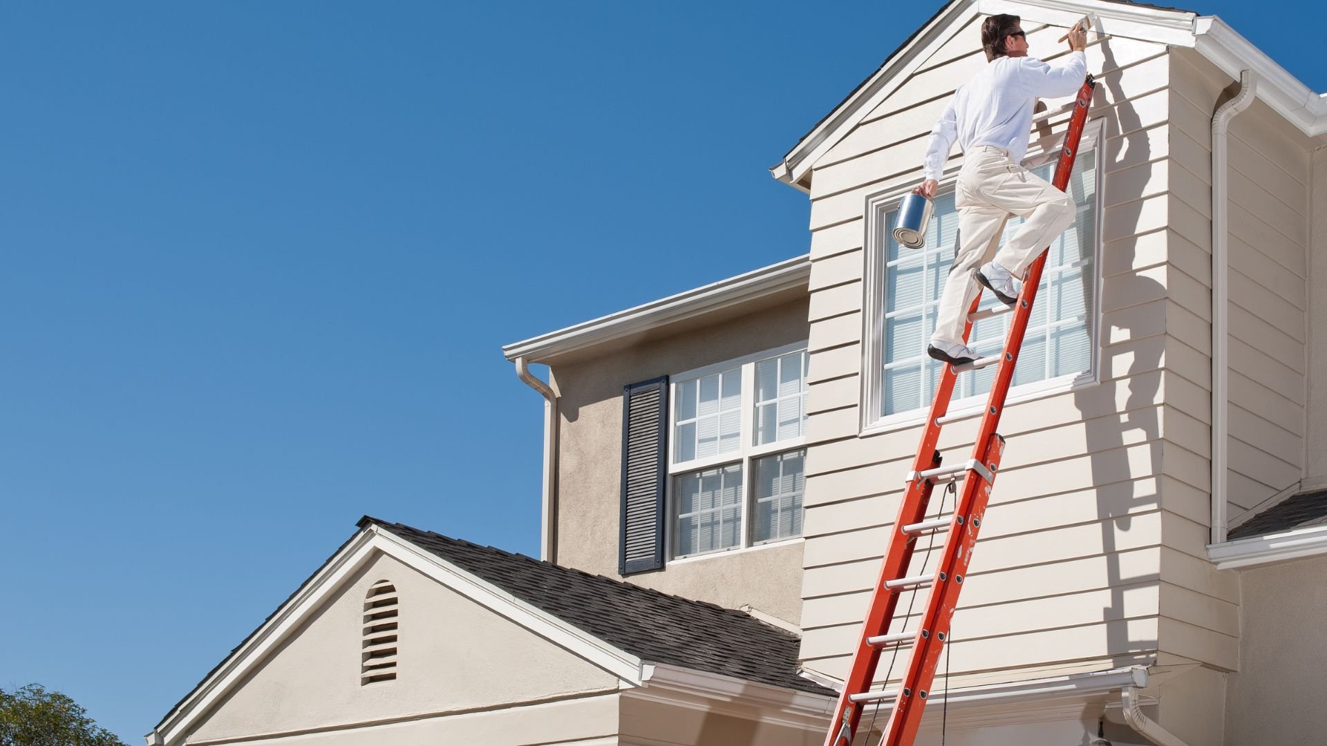 Painter on a red ladder working on the exterior of a white two-story house
